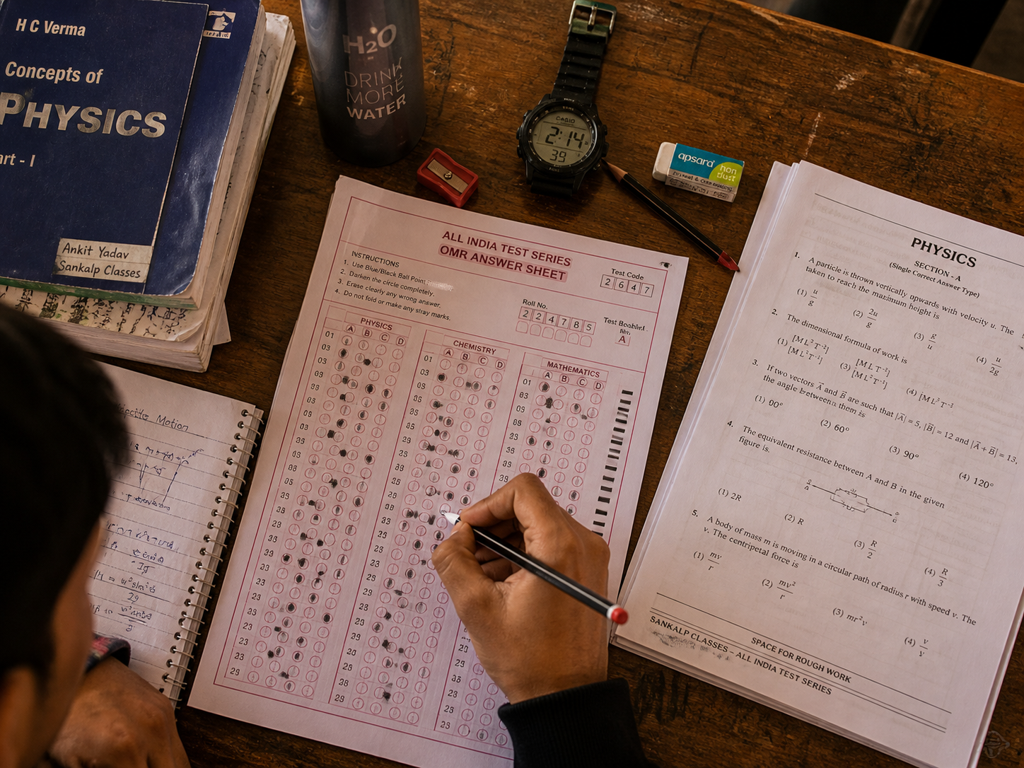 Sankalp Classes test series — top-down student desk during JEE-pattern mock test, OMR answer sheet half-filled, physics paper, watch, water bottle, eraser